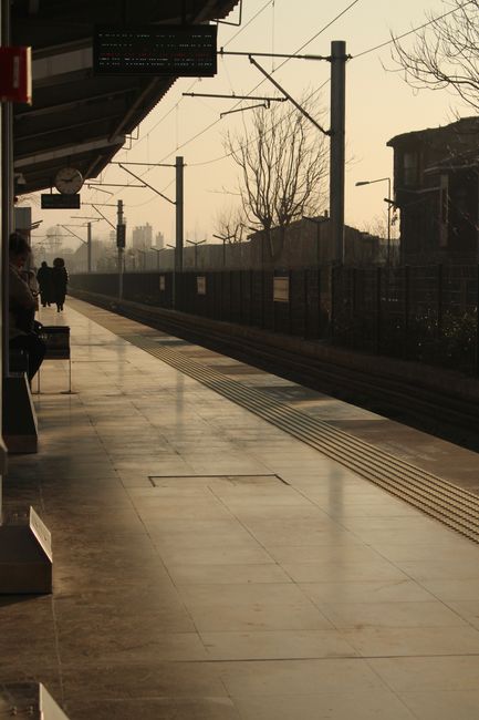 A quiet train platform at Pinner Station during early evening, with a polished concrete surface showing reflections of the overhead lighting. The platform features tactile paving strips near the edge 
