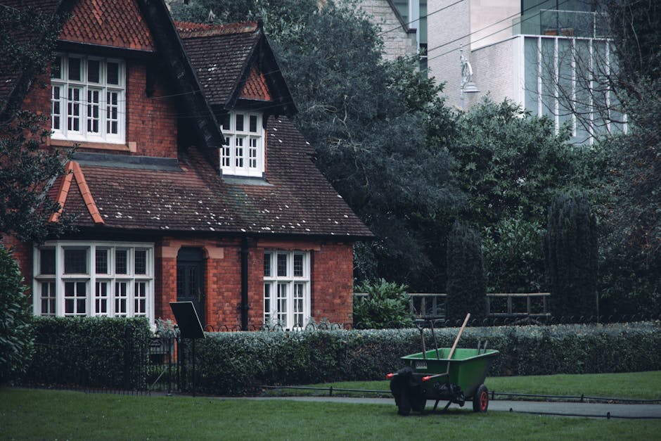A red brick house with a steep, tiled roof features white-framed sash windows and a black front door, surrounded by a well-maintained garden. In the foreground, a green wheelbarrow with red wheels and