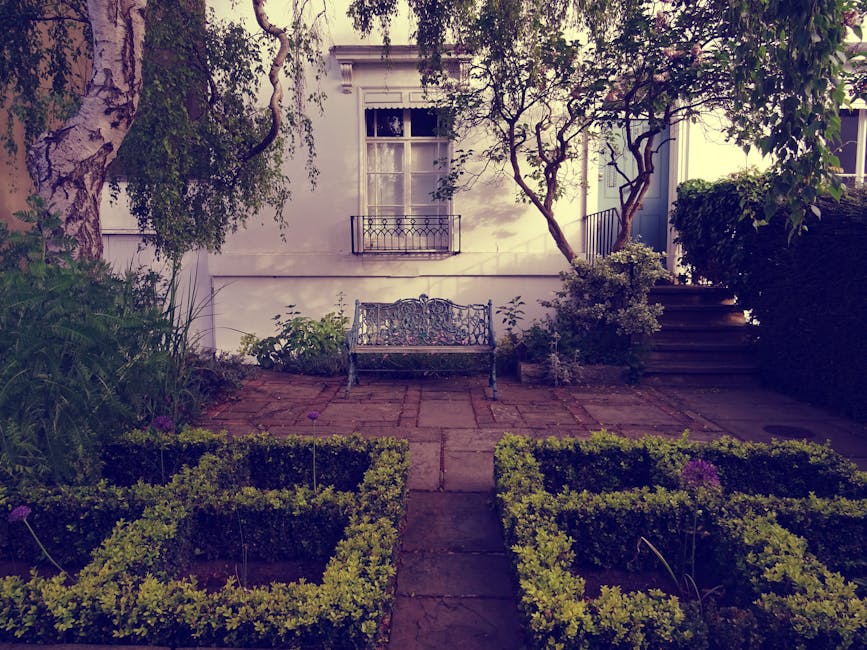 A red brick house with a steep, tiled roof features white-framed sash windows and a black front door, surrounded by a well-maintained garden. In the foreground, a green wheelbarrow with red wheels and